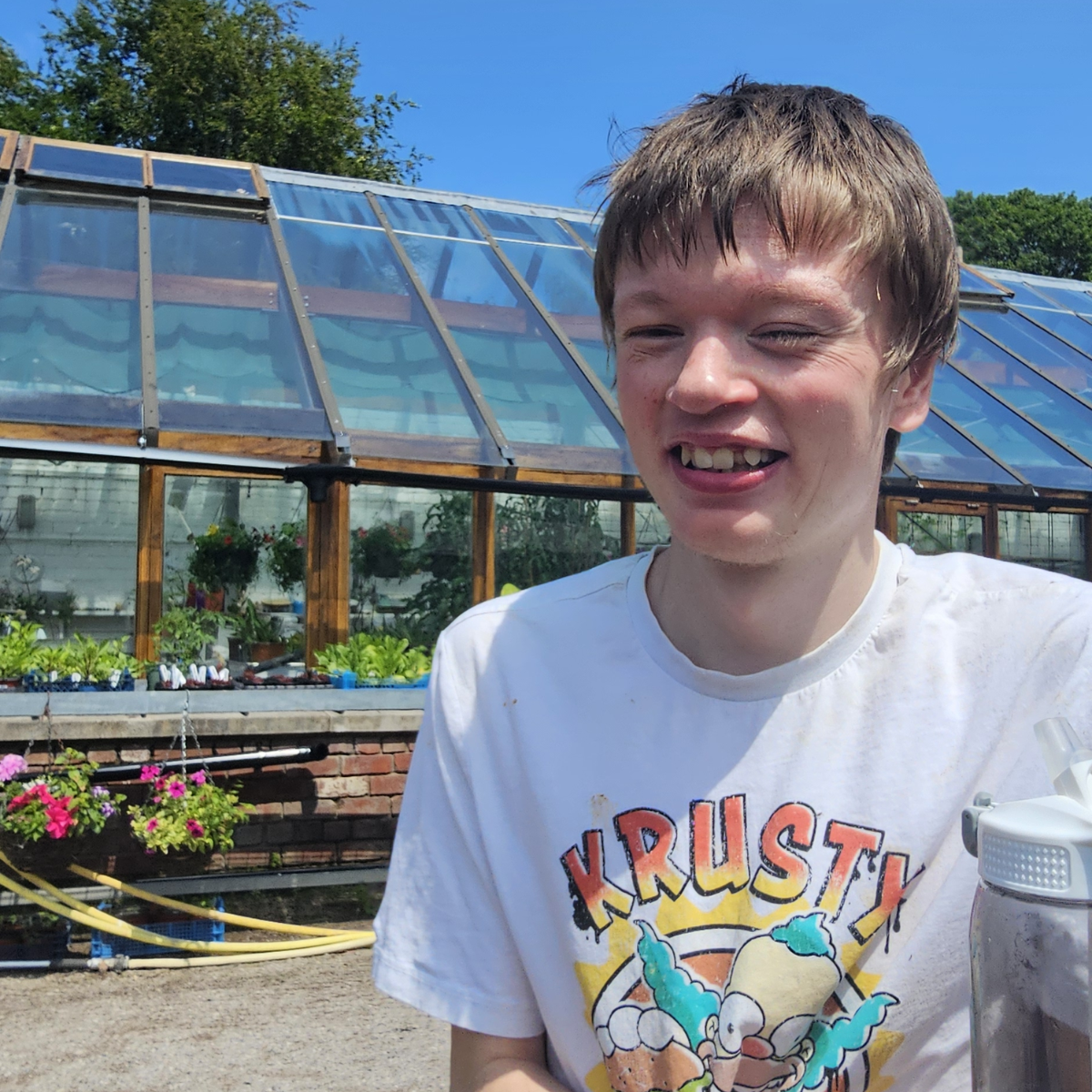 Young man in a Krusty Burger t-shirt smiles in front of a greenhouse on a sunny day. Plants and flowers are visible.