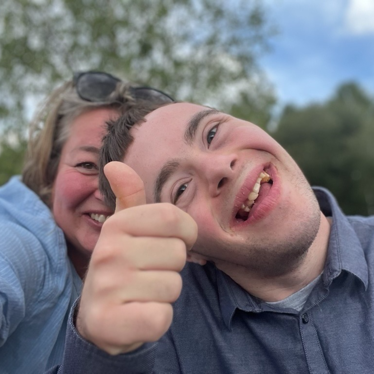 A woman and a young man with Down syndrome smile for a selfie outdoors, with the man giving a thumbs up.