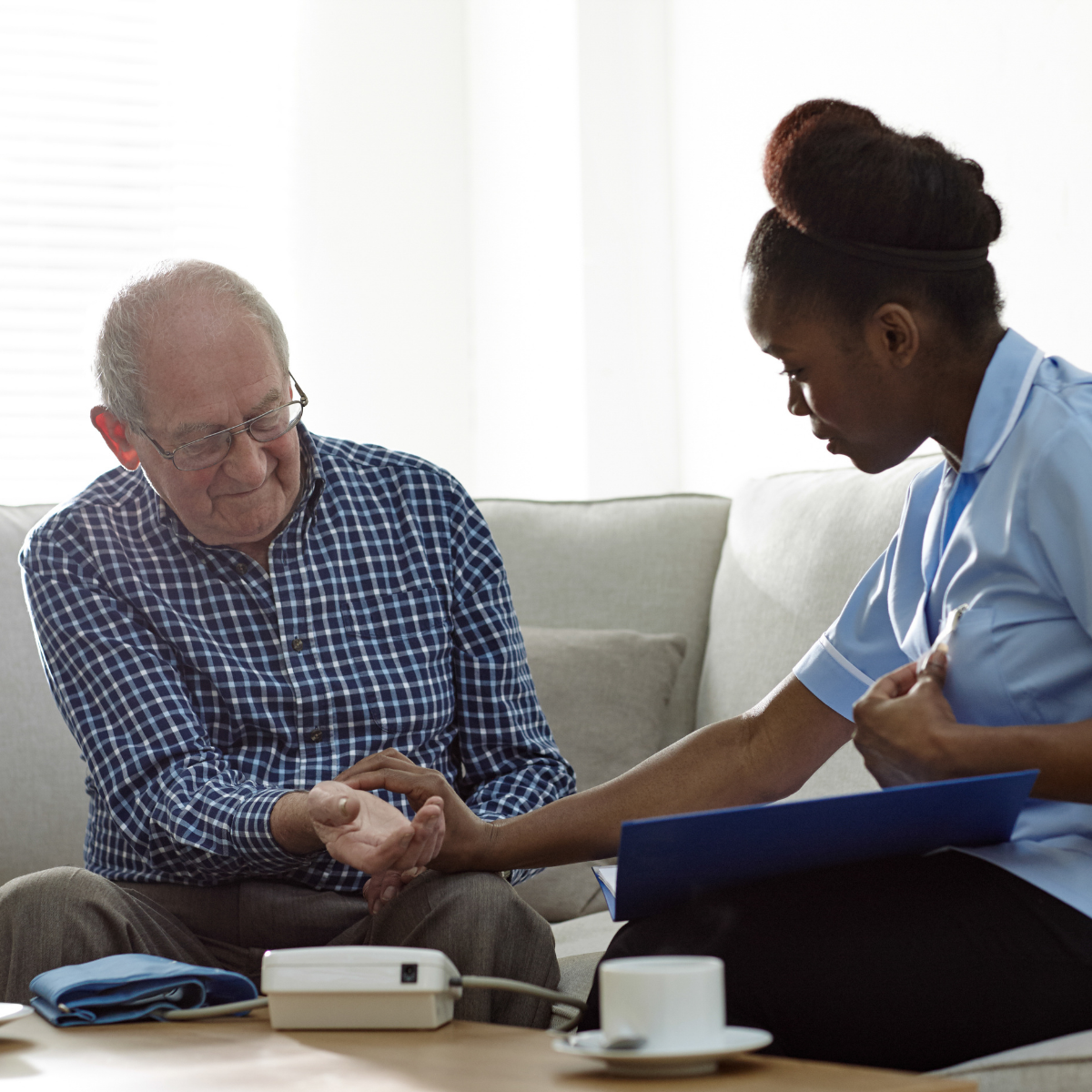 A nurse checks an elderly man's pulse at his home, with a blood pressure monitor nearby.