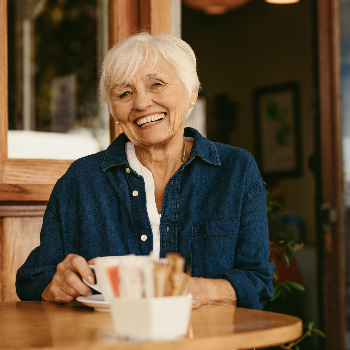 A happy senior woman with white hair and a denim shirt smiles broadly while holding a coffee cup at an outdoor cafe.