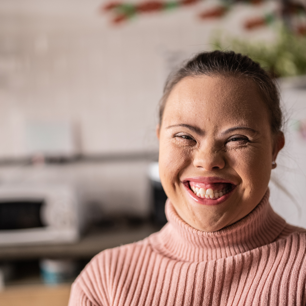 A young woman with Down syndrome smiles widely, wearing a pink turtleneck sweater.