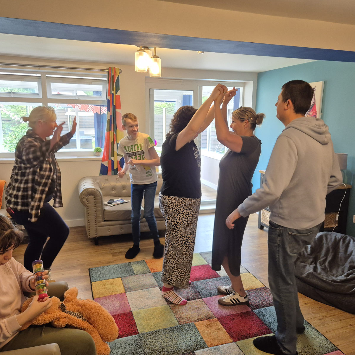 A group of people, including children and adults, are dancing and holding hands in a brightly lit living room.