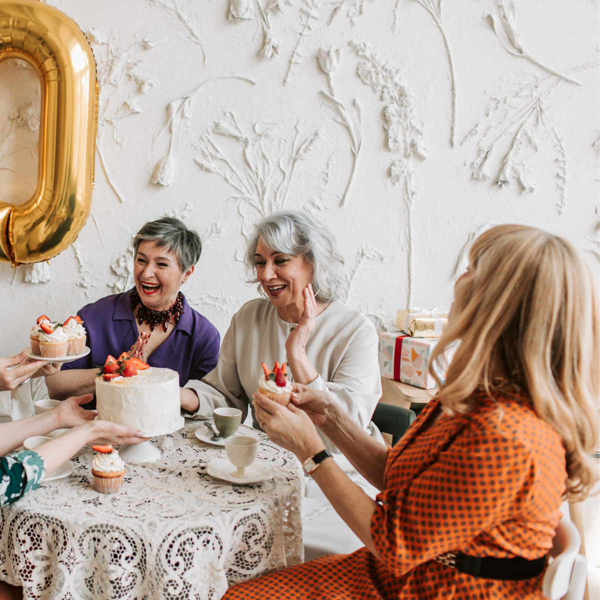 Women celebrating a birthday with cake and cupcakes, with a large gold "0" balloon.