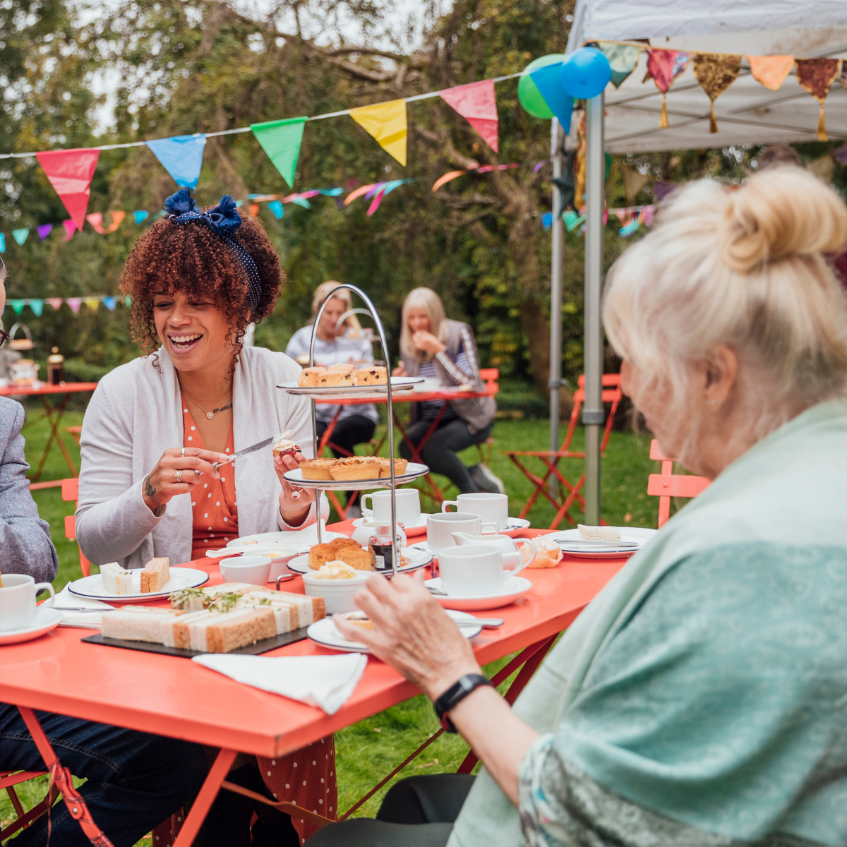 A group of women enjoy afternoon tea outdoors, with colorful banners and balloons decorating the scene.